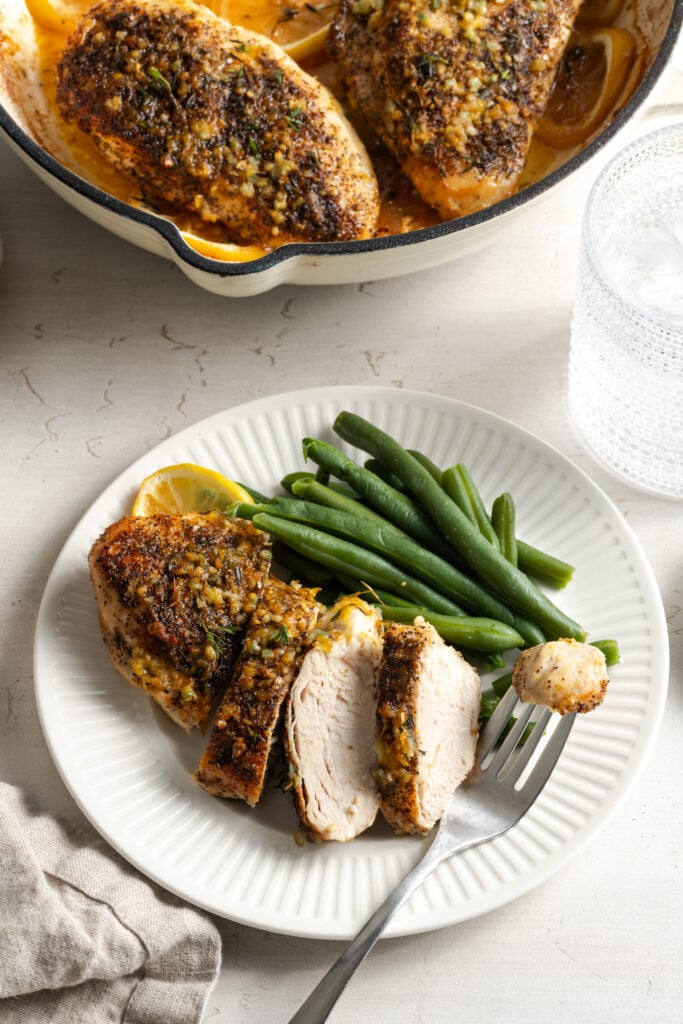 sliced lemon pepper chicken breast on a white dinner plate with green beans; a bite is on a silver fork and there's a glass of water on the table.