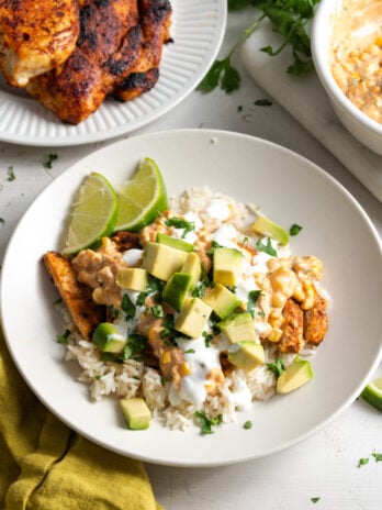 white work surface with herbs scattered around after cooking with an assembled street corn and chicken bowl next to a plate with the remaining chicken and a bowl with the remaining elote.