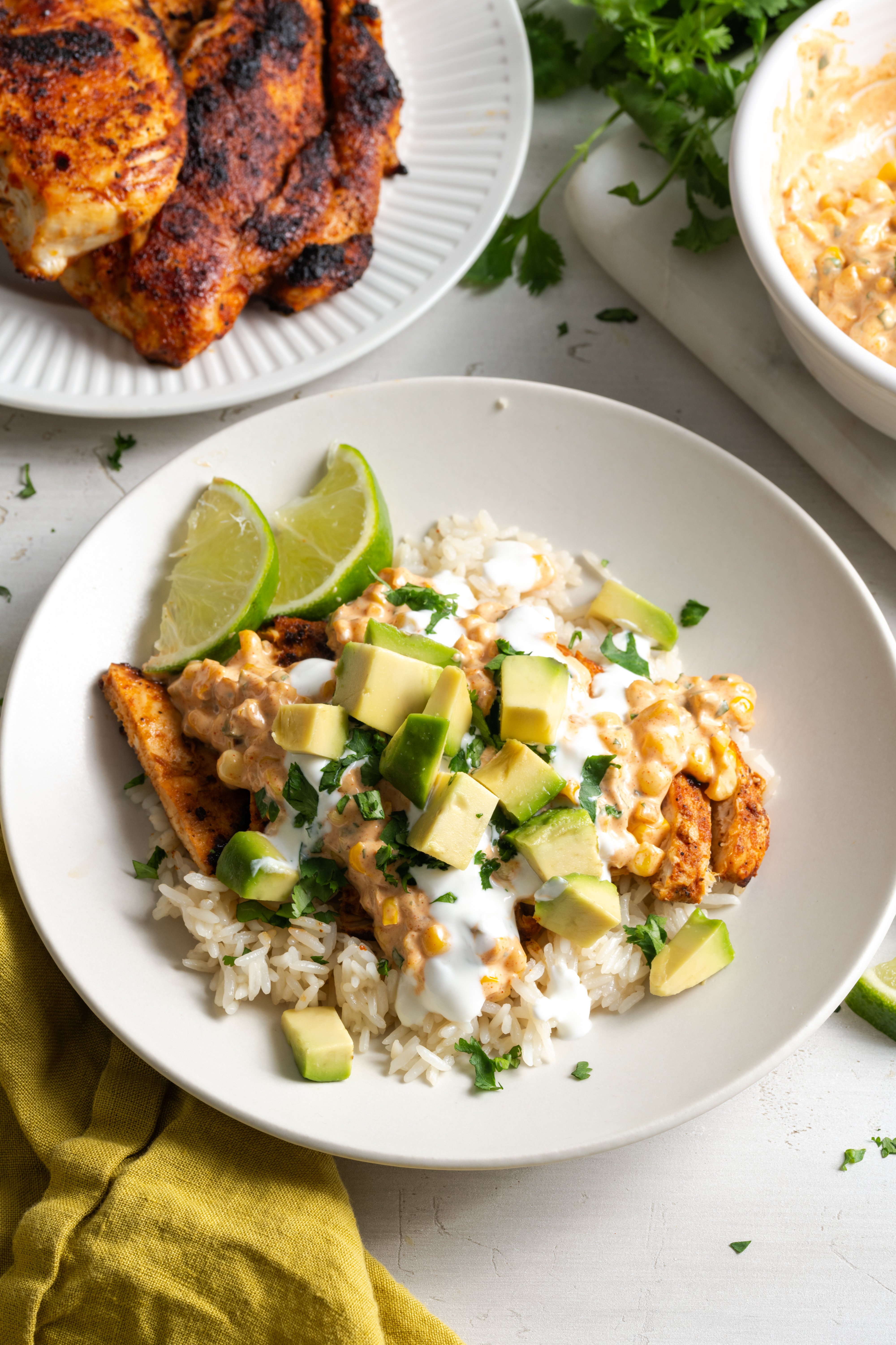 white work surface with herbs scattered around after cooking with an assembled street corn and chicken bowl next to a plate with the remaining chicken and a bowl with the remaining elote.