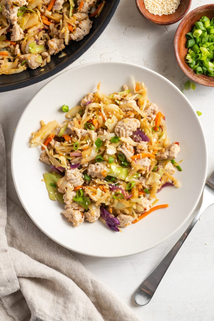 white pasta bowl of ground turkey egg roll in a bowl on a white table with a silver fork, a pinch bowl of scallions and a pinch bowl of sesame seeds, with a white cloth napkin.