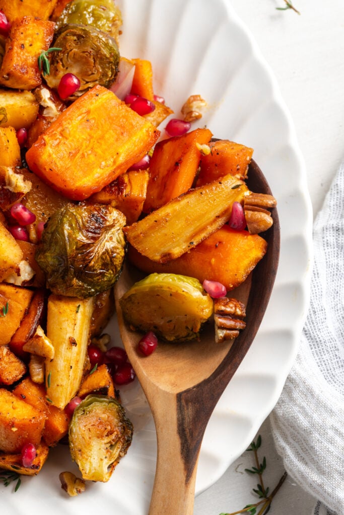 overhead shot of fall roasted vegetables on a white serving platter with a wooden serving spoon taking a scoop.