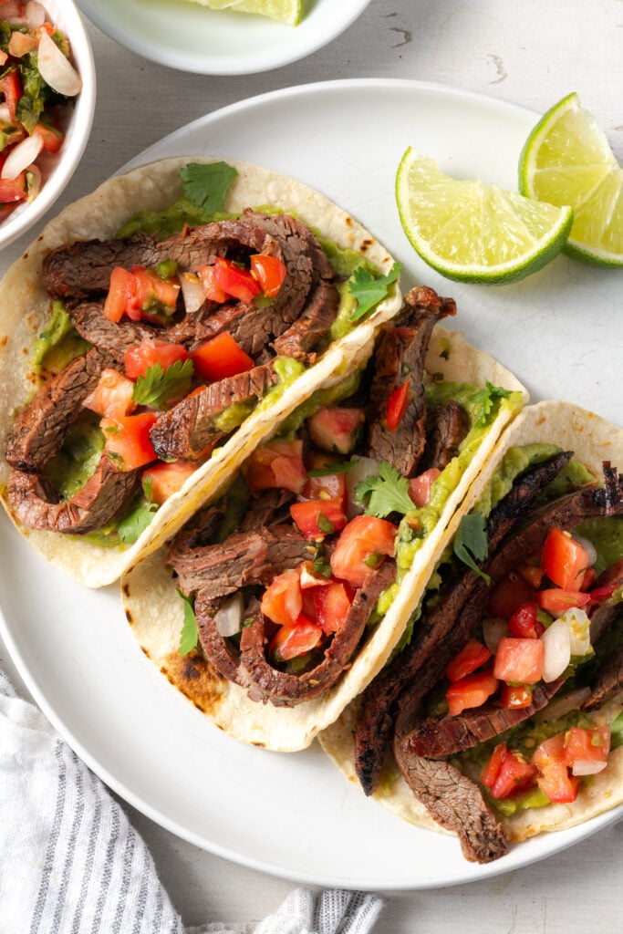 white plate with three flank steak tacos with two lime wedges.