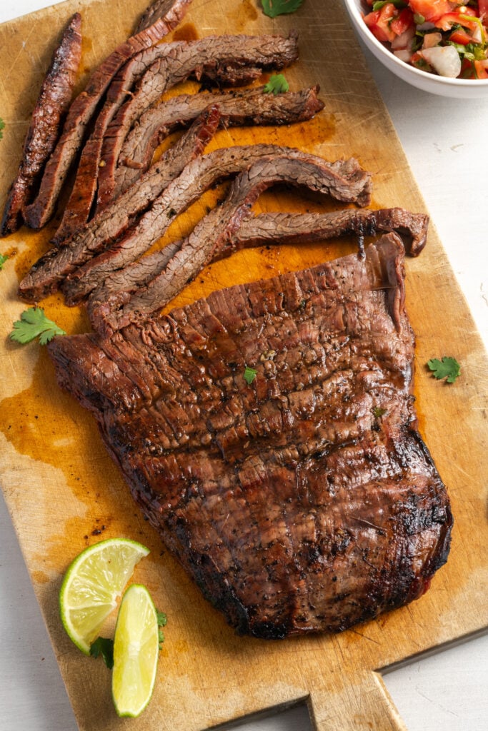 marinated flank steak on a wooden cutting board set aside for tacos after slicing with a bowl of pico de gallo and fresh cilantro sprinkled over.