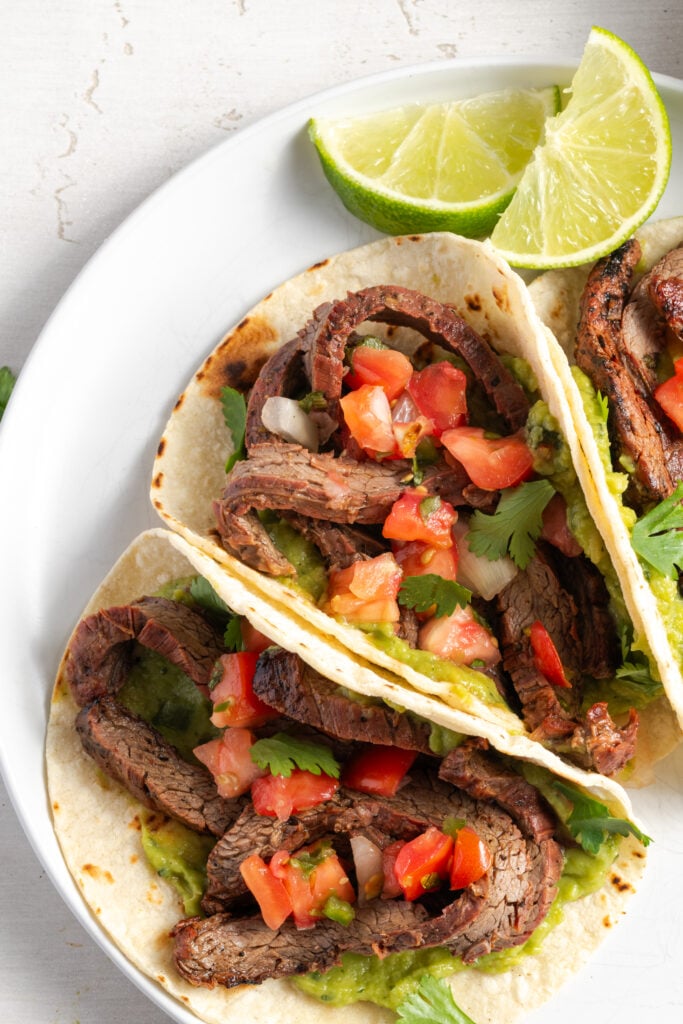 sliced marinated flank steak tucked into tacos with avocado mash, pico de gallo, and fresh cilantro on a white plate with lime wedges.