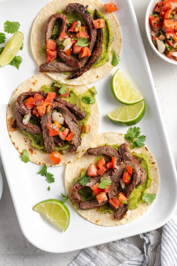 white rectangular serving platter with three flank steak tacos next to a bowl of pico de gallo with sliced limes.