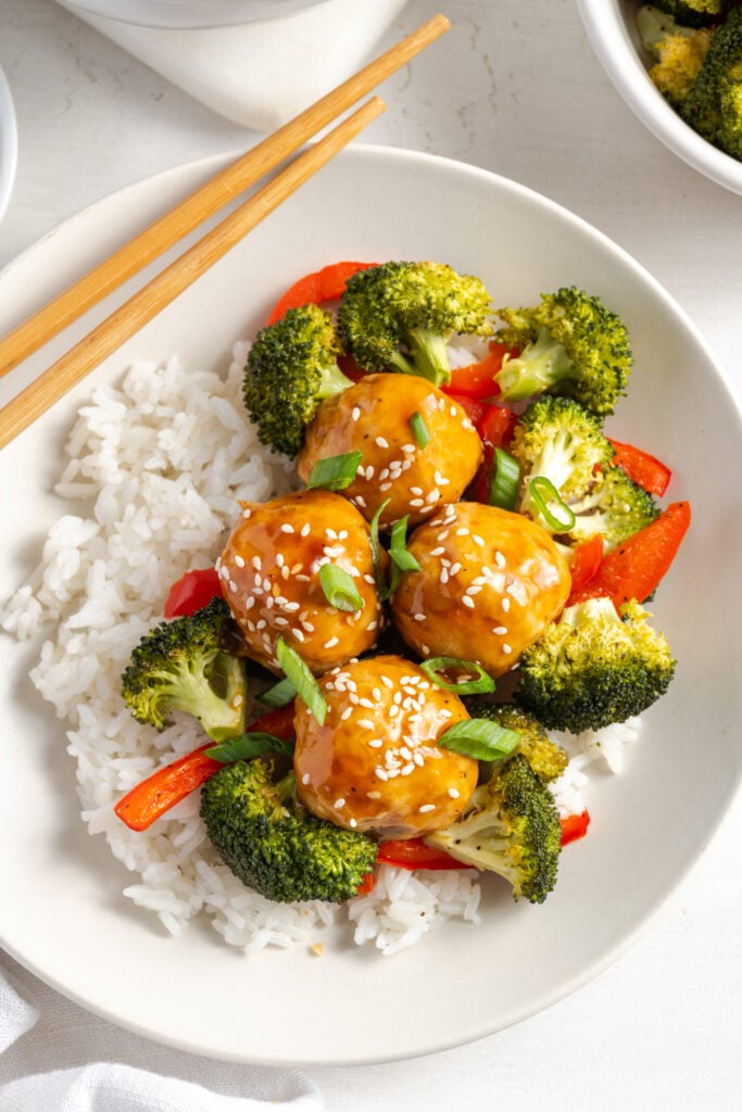 plate of chicken meatballs with roasted veggies tossed in a pineapple teriyaki sauce over a bed of steamed white rice with a pair of chopsticks.