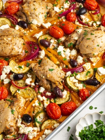 greek sheet pan chicken thighs and veggies on the sheet pan on a white table with a small white pinch bowl of fresh parsley.