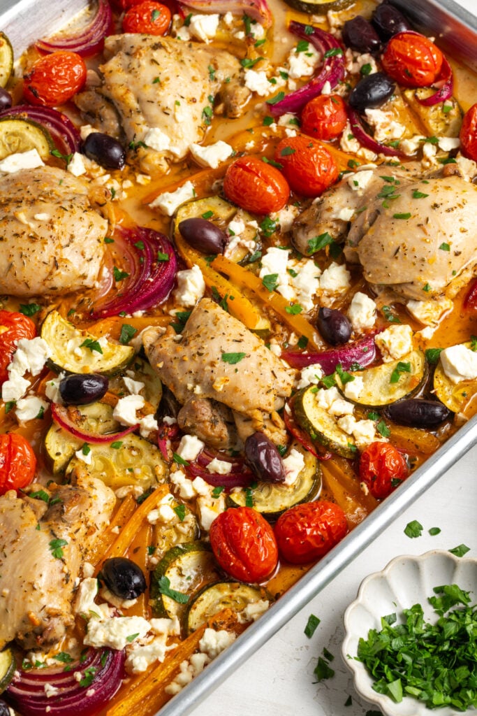 greek sheet pan chicken thighs and veggies on the sheet pan on a white table with a small white pinch bowl of fresh parsley.