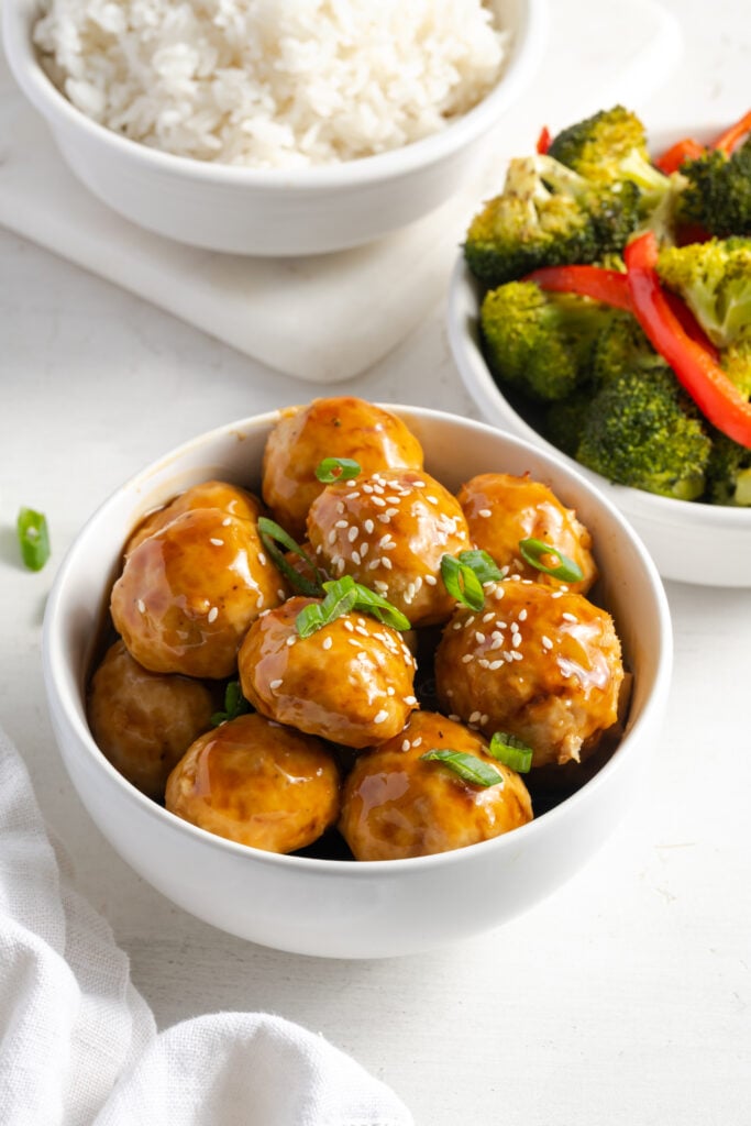 pineapple teriyaki chicken meatballs in a white bowl on a white table with bowls of rice and roasted veggies.