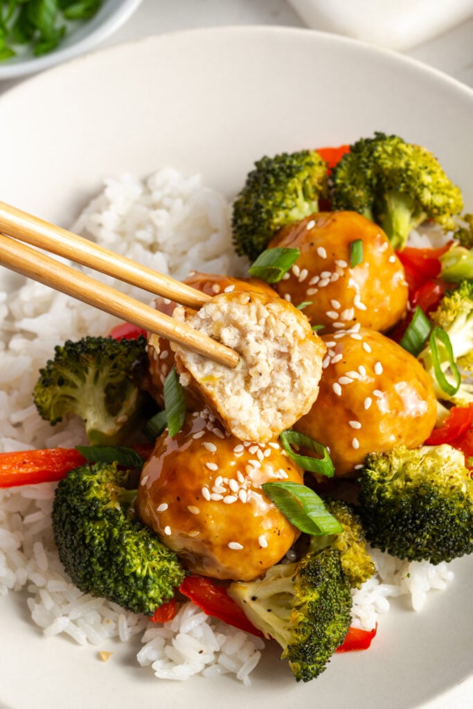 wooden chopsticks holding a pineapple teriyaki ground chicken meatball with a bite taken out of it above a bowl filled with rice, roasted veggies, and more meatballs.
