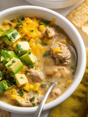 white soup bowl filled with ground turkey white chili topped with cheddar cheese, avocado cubes, and fresh cilantro on a white table with a saffron yellow napkin.
