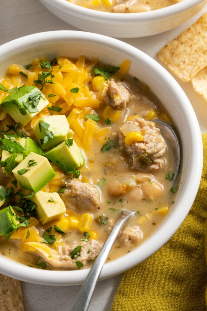 white soup bowl filled with ground turkey white chili topped with cheddar cheese, avocado cubes, and fresh cilantro on a white table with a saffron yellow napkin.