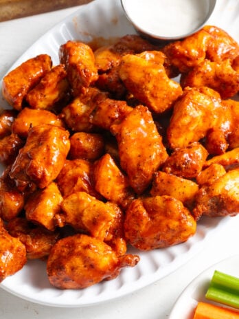 45 degree angle shot of gluten-free buffalo chicken bites on a white serving platter with a ramekin of ranch dressing and some carrot and celery sticks on a separate white plate.