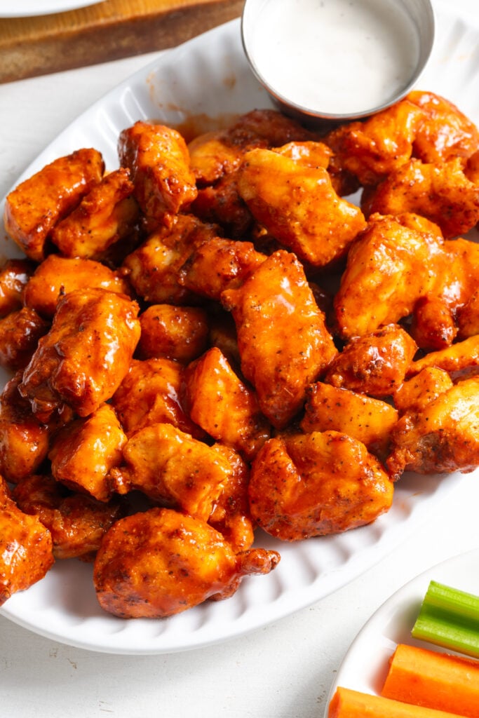 45 degree angle shot of gluten-free buffalo chicken bites on a white serving platter with a ramekin of ranch dressing and some carrot and celery sticks on a separate white plate.