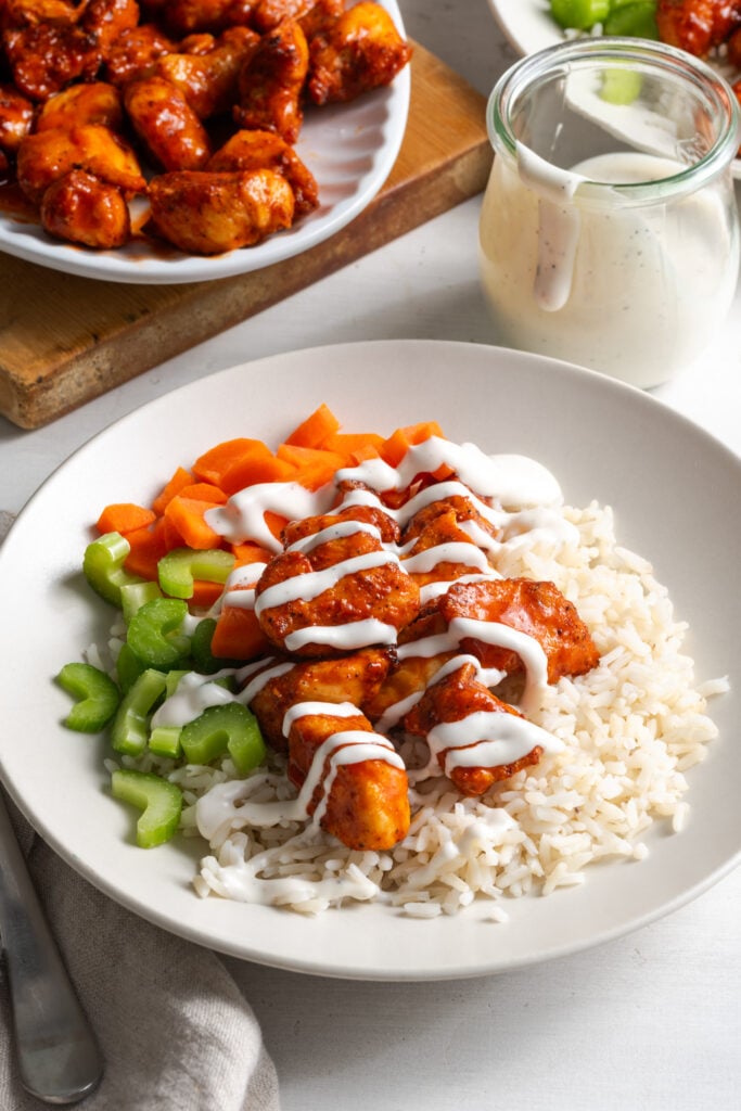 dinner table with a white pasta bowl filled with steamed white rice topped with air fryer buffalo chicken bites, chopped celery and carrots, and a drizzle of ranch; a small glass jar with extra ranch and the serving platter with the rest of the bites is in the top of the frame.