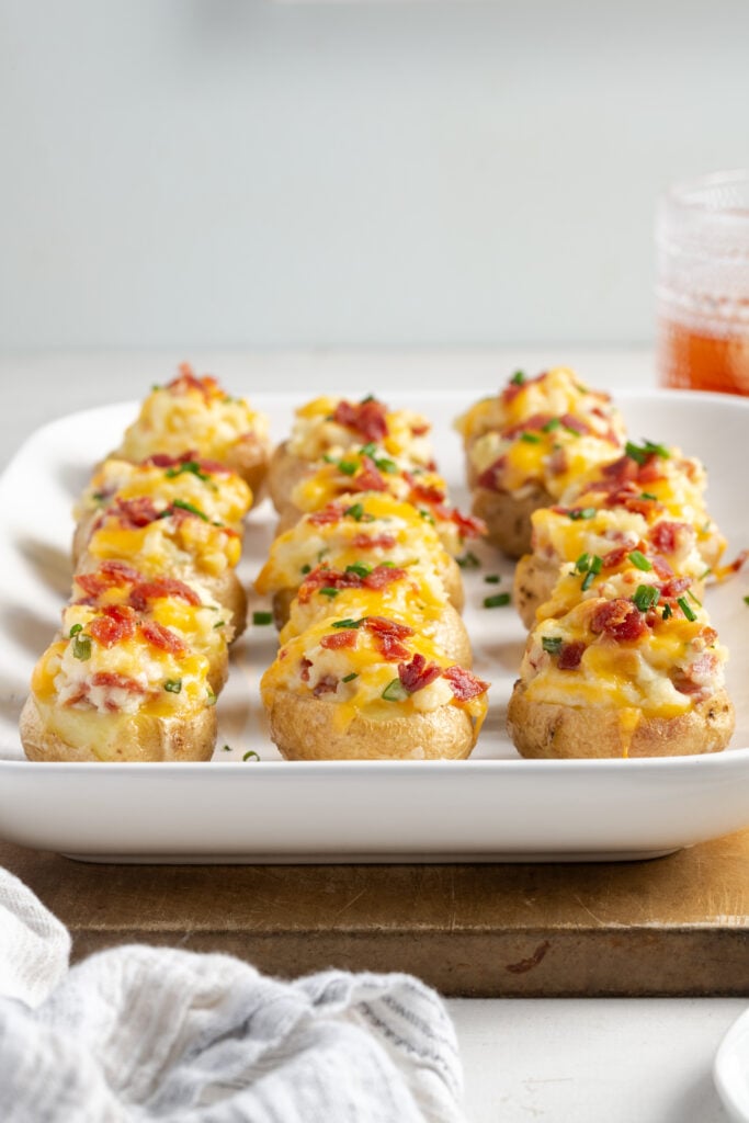 side on shot of a serving tray of loaded twice baked baby potatoes on a wooden cutting board on a white table with a white background.