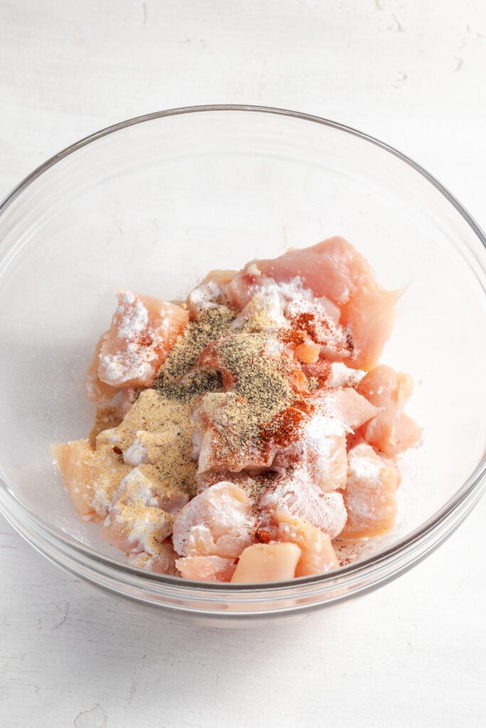 raw chicken bites in a mixing bowl with seasonings on top.