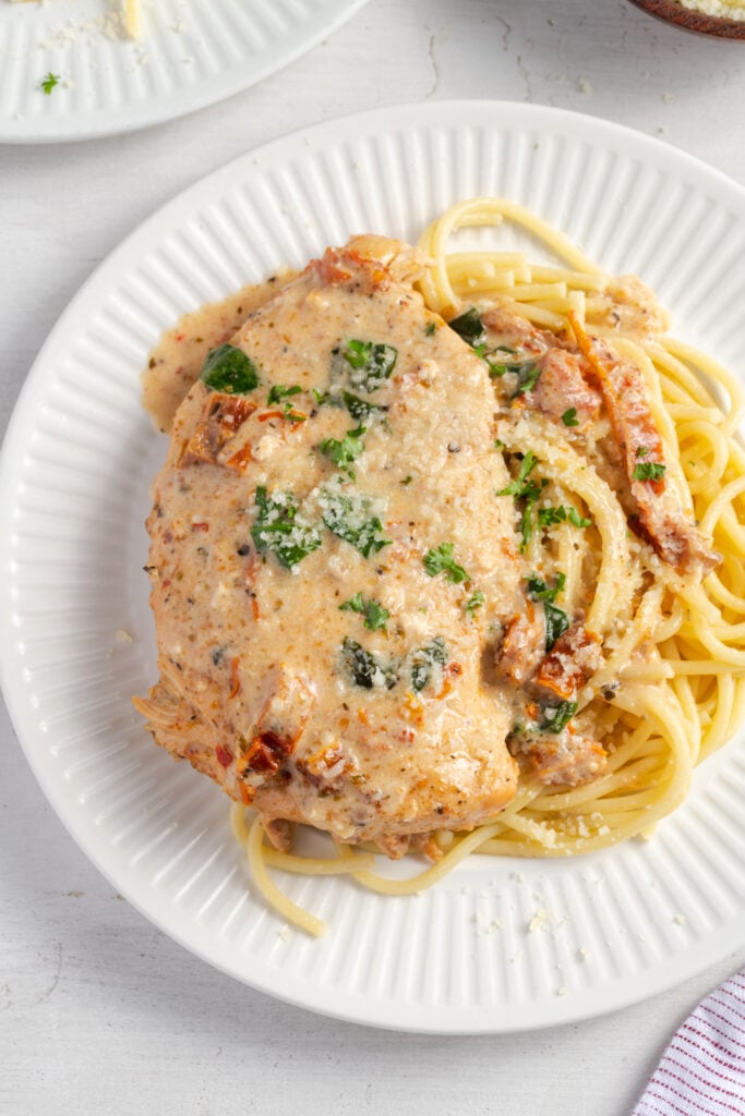 overhead shot of a plate of slow cooker tuscan chicken breast on a bed of cooked pasta on a white plate on a white table.