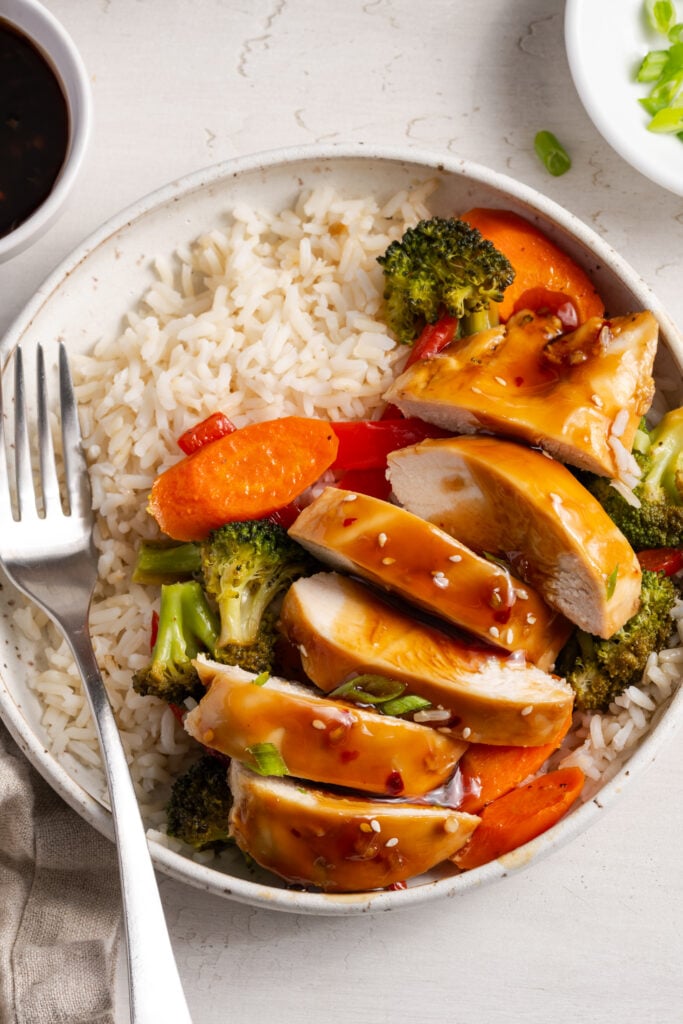 overhead shot of easy baked chicken teriyaki with carrots and broccoli served on a bed of rice in a white pasta bowl with a silver fork.
