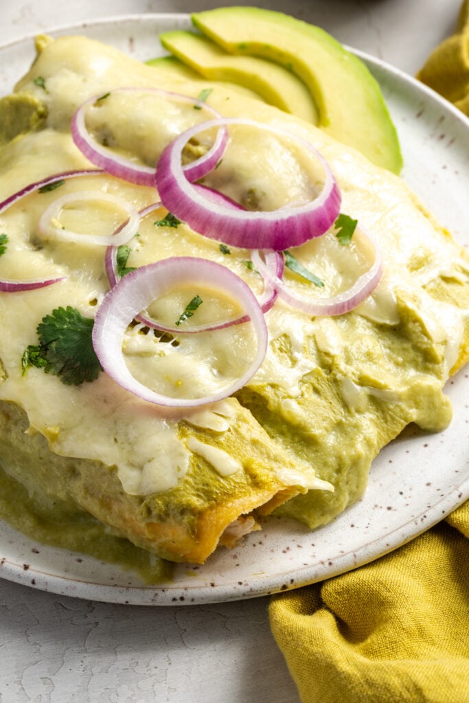 white earthenware plate with 3 green chili chicken enchiladas garnished with red onion rings and avocado slices on a grey table with a yellow napkin.