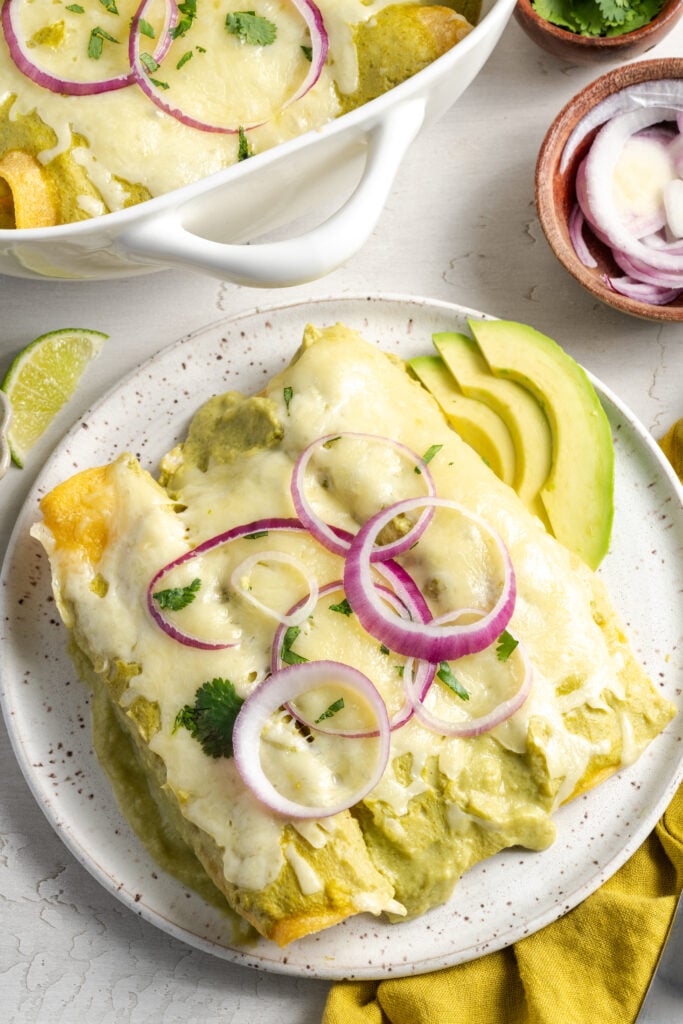 serving of green chicken enchiladas on a plate with fresh garnishes on a white table with a yellow napkin and a slice of lime.