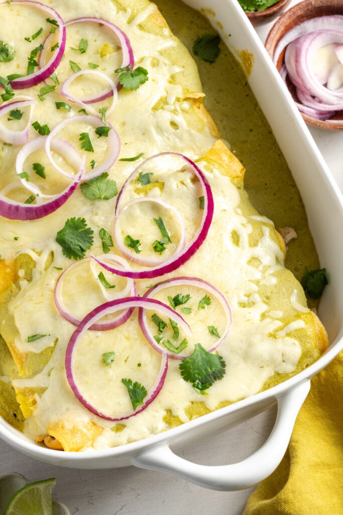 overhead shot of the bottom 2/3 of a casserole dish filled with green chicken and cheese enchiladas topped with sliced red onions and fresh cilantro leaves.
