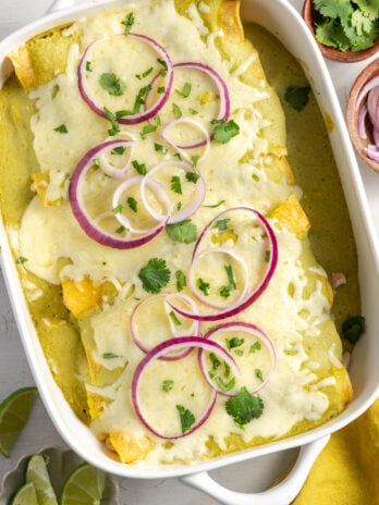 flat lay shot of a white casserole dish with green chile chicken enchiladas topped with fresh cilantro and red onion ring slices on a white table with pinch bowls of lime slices, more onions, and more cilantro.