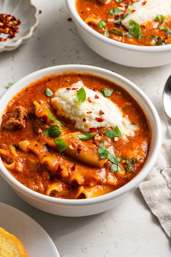 45 degree angle showing two white bowls filled with slow cooker lasagna soup garnished with extra ricotta, fresh basil, and red pepper flakes.