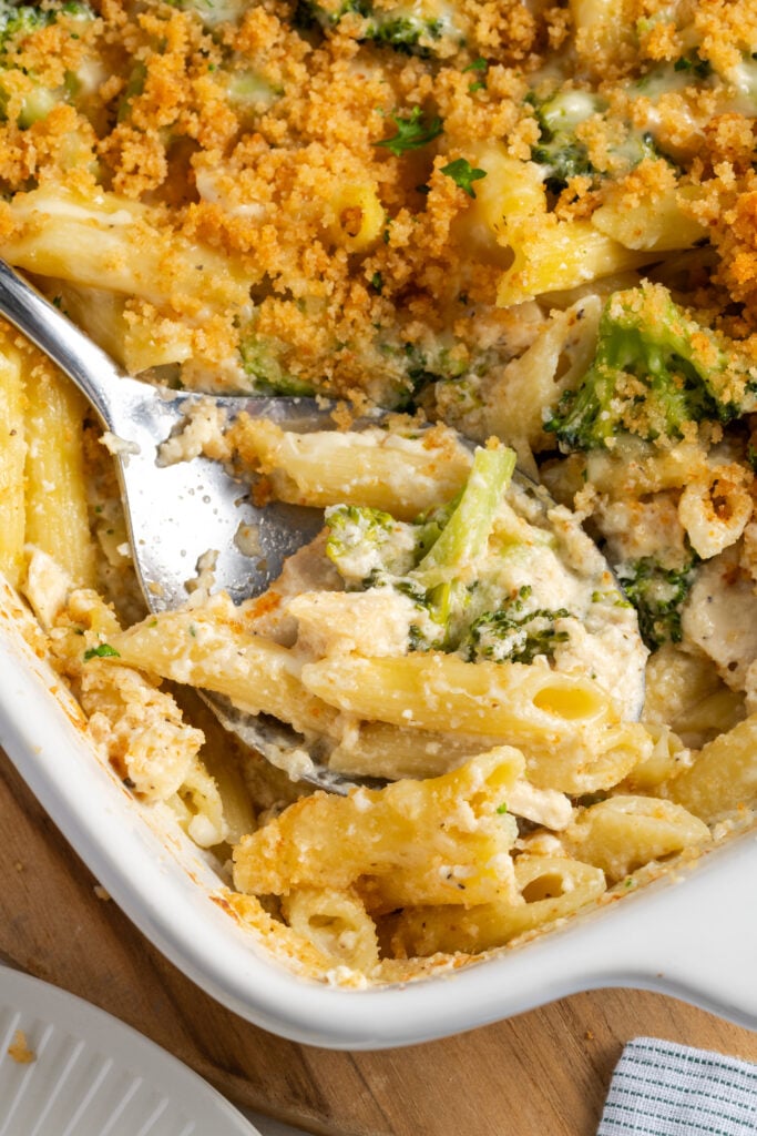 closeup shot of the corner of the baking dish with a silver serving spoon dishing out some of the creamy broccoli chicken alfredo, showing the contrast with the crunchy breadcrumbs on top.