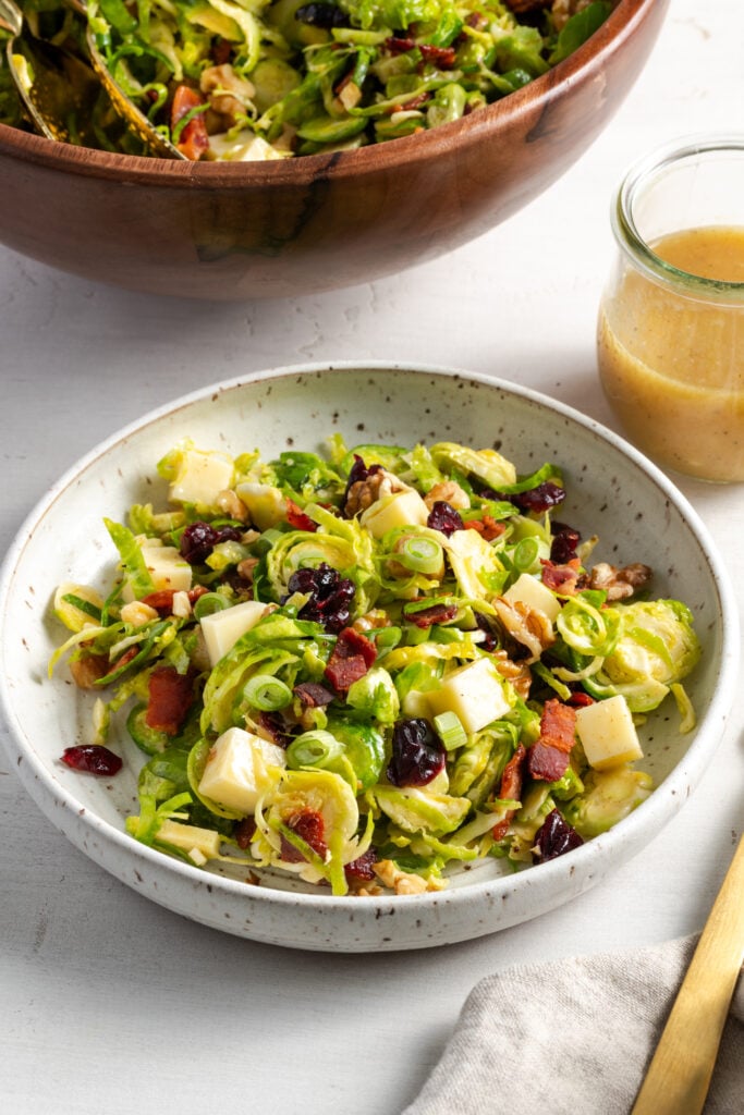 45 degree angle shot of a white flecked earthenware shallow bowl filled with a serving of shaved brussels sprouts salad on a white table with the wooden serving bowl and a small glass jar with extra vinaigrette.