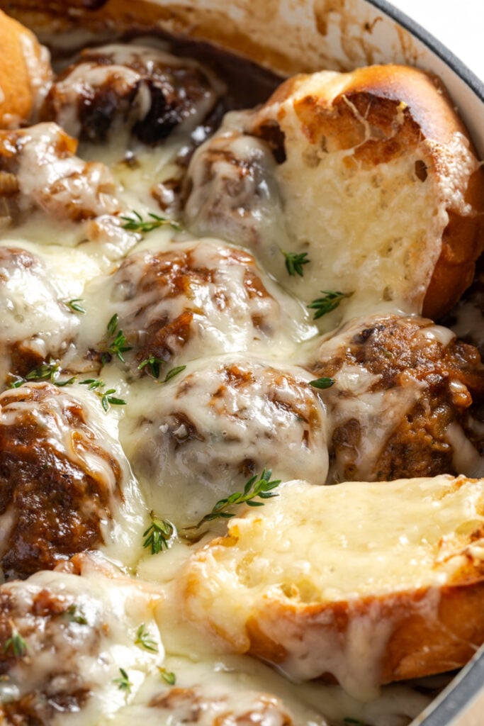 closeup 45 degree angle shot of baked french onion beef meatballs topped with melted cheese and slices of french onion sticking out of the gravy in the baking dish.