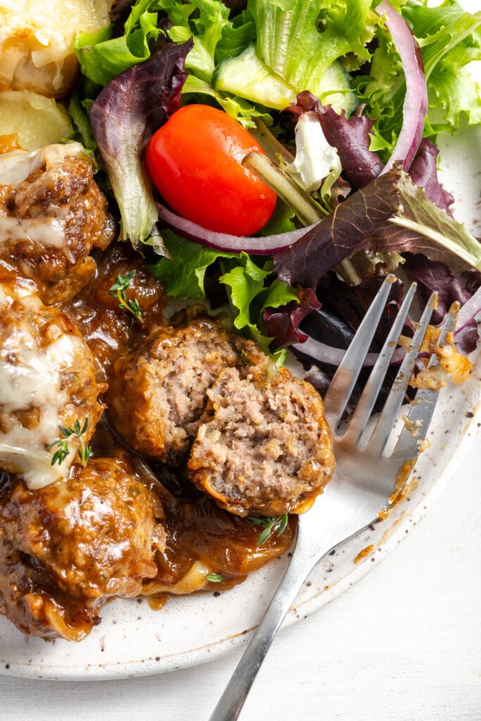 closeup overhead shot of a french onion meatball sliced in half on the plate with the silver fork to show the tender ground beef interior, thick onion gravy, and a bit of the side salad.