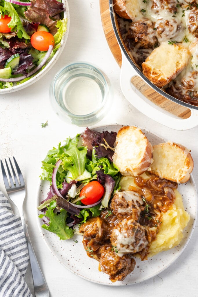 overhead shot of a white table with a serving of french onion meatballs served over creamy mashed potatoes with a side salad on a white earthenware plate; the table also has a silver fork, a grey and white striped napking, a salad bowl with more salad, and the corner of the baking dish with the rest of the meatballs and bread in it.