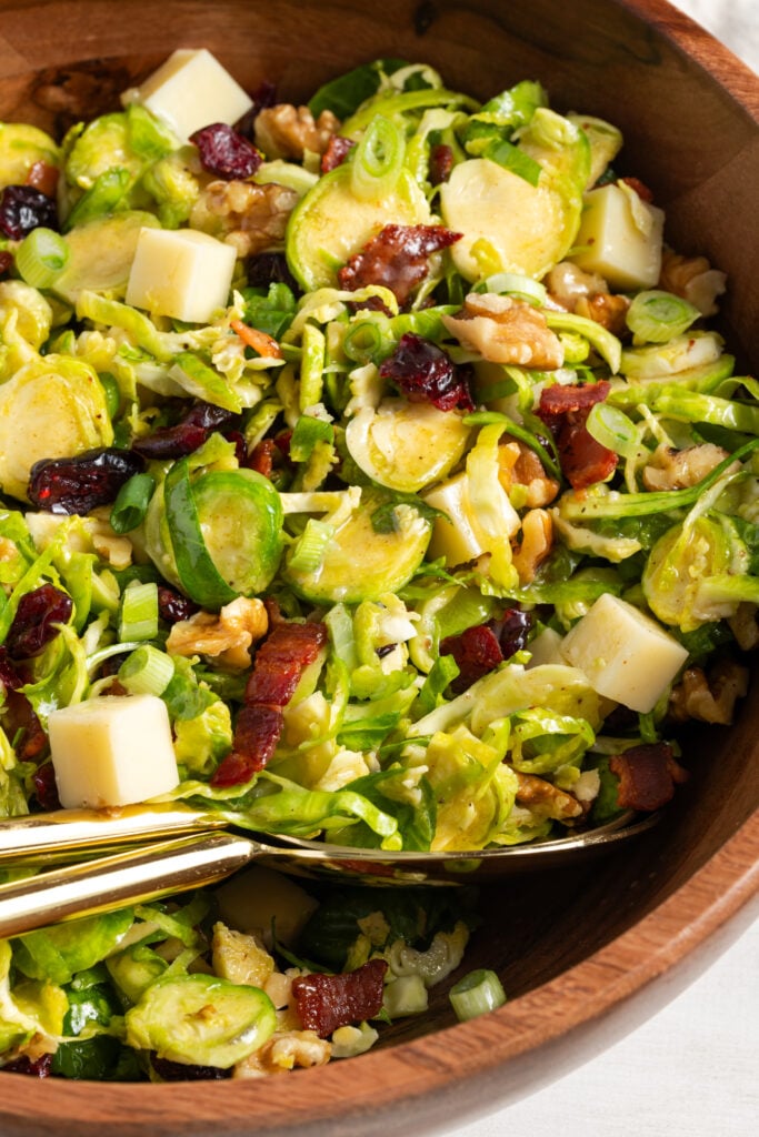 closeup of shaved brussel sprout salad in a wooden serving bowl with gold serving spoons.