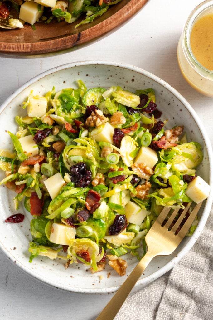 overhead shot of a flecked white earthenware pasta bowl filled with a portion of shaved brussel sprouts salad with bacon, cheese, and cranberries on a white table with a gold fork.