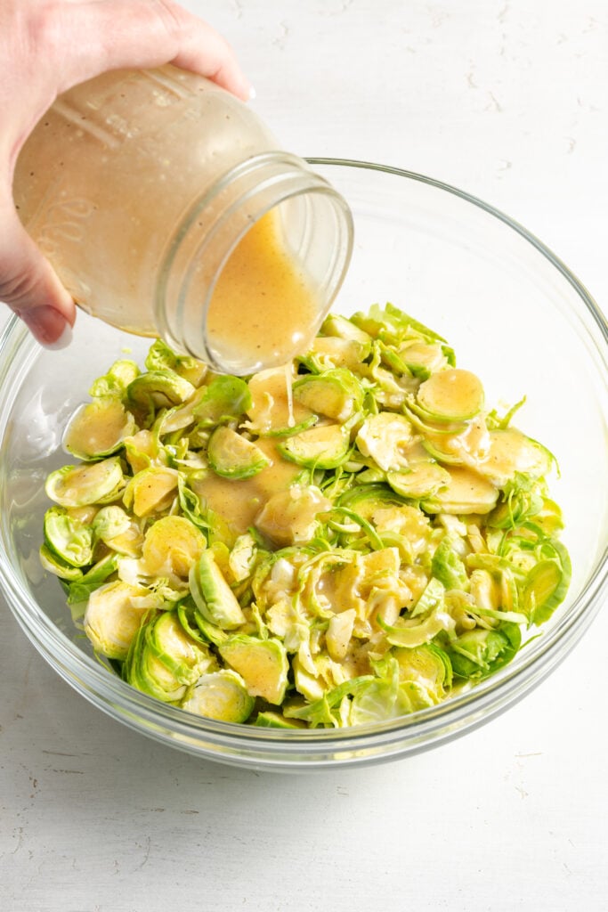 hand pouring vinaigrette from a mason jar over the shaved brussels sprouts in the bowl.