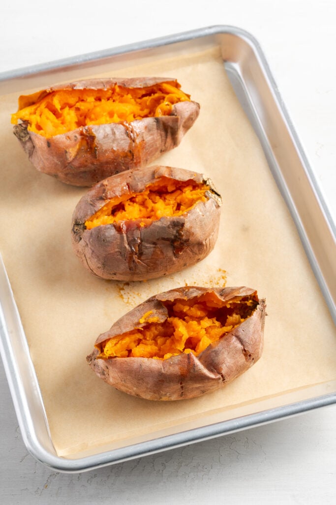 three baked sweet potatoes on a sheet pan after slicing a pocket and fluffing the insides a bit.