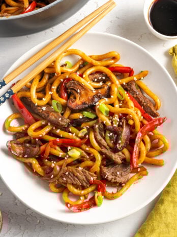 white pasta bowl of beef and noodle stir-fry on a white table with a yellow napkin and wooden chopsticks.