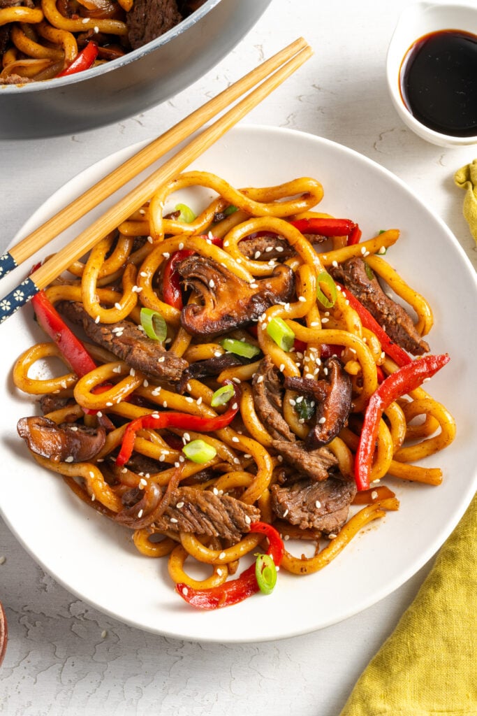 white pasta bowl of beef and noodle stir-fry on a white table with a yellow napkin and wooden chopsticks.