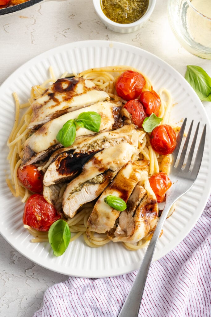 overhead shot of a dinner plate with sliced caprese chicken drizzled with balsamic reduction and topped with fresh basil on a white table with a silver fork and a glass of white wine.