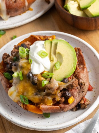 ground beef and black bean stuffed sweet potato on a white plate with avocado slices, a dollop of sour cream, and fresh green onions.