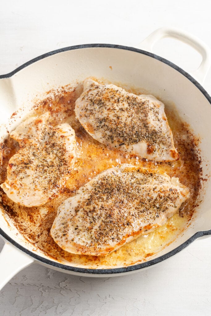 chicken breasts searing in a white enamel cast iron skillet.