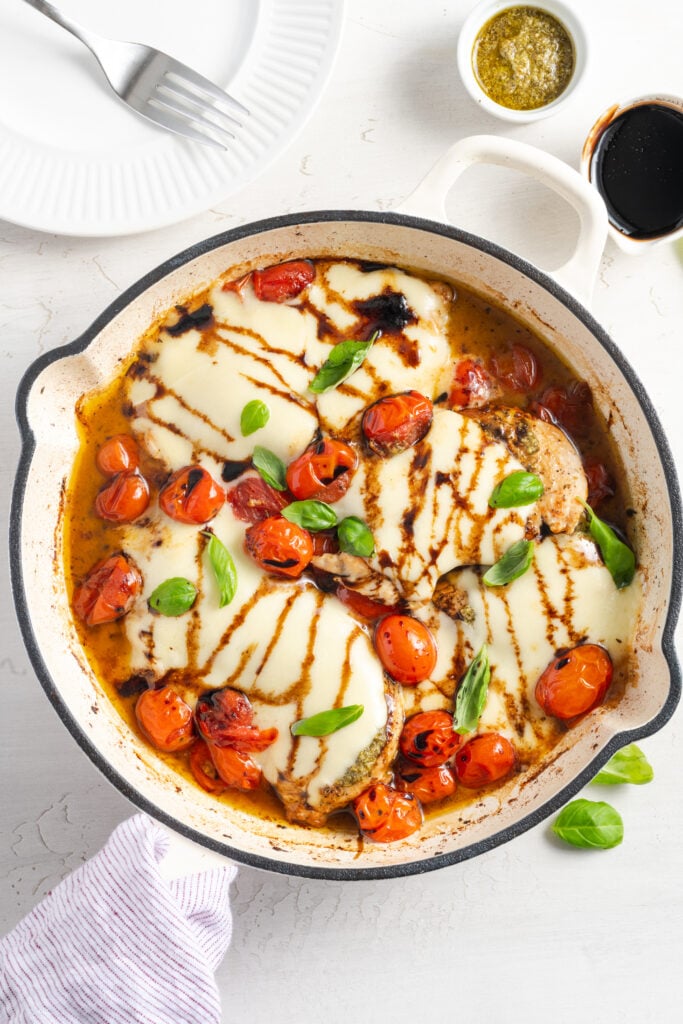 skillet chicken caprese in a white ceramic-coated skillet topped with fresh basil and balsamic reduction on a white table with a white plate, silver fork, and a small bowl of extra pesto.