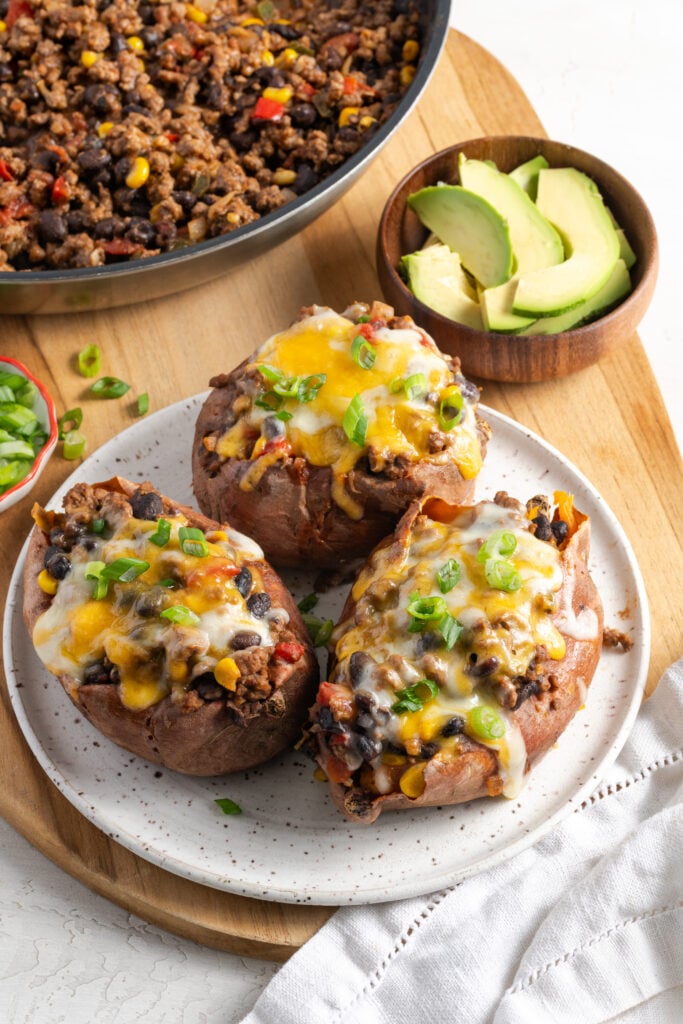 southwestern ground beef stuffed sweet potatoes on a plate topped with melted cheese and fresh scallions with the skillet of ground beef and black bean taco filling and a bowl of sliced avocados in the top of the frame.