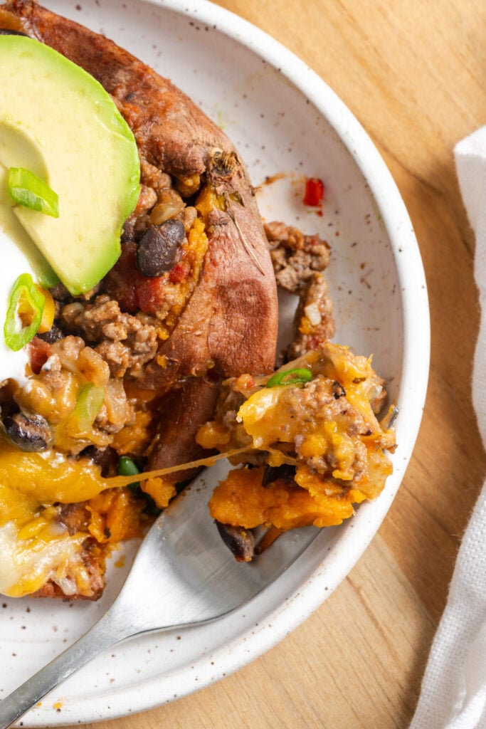 closeup of a silver fork taking a bite of a stuffed baked sweet potato showing the stretchy, melty cheese and hearty black bean and ground beef taco filling.