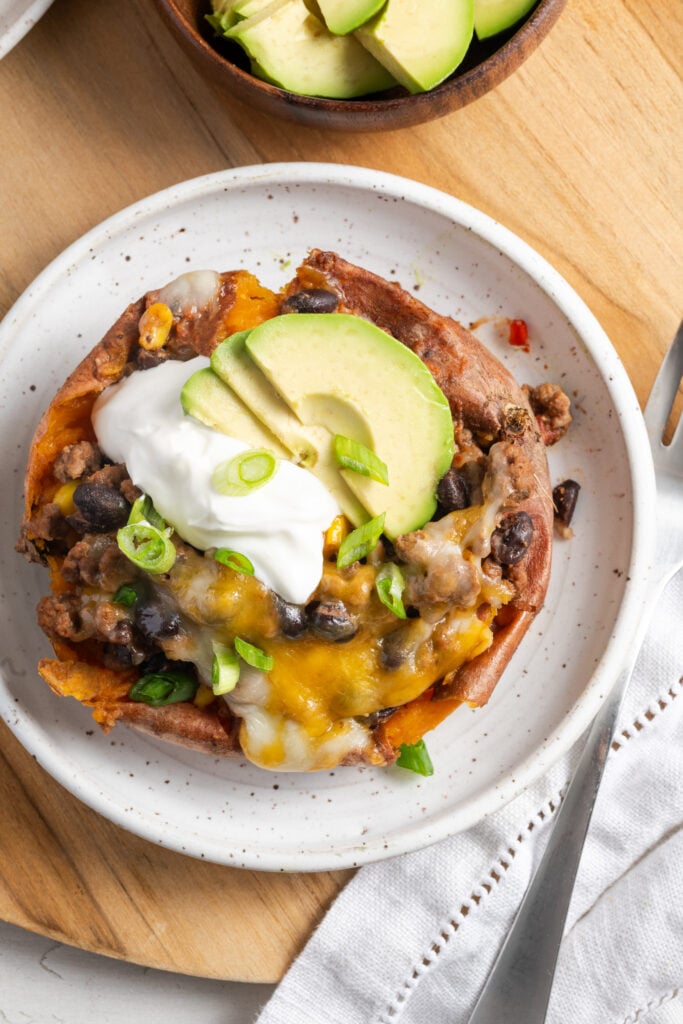 overhead closeup shot of a taco stuffed sweet potato garnished with sour cream, avocado slices, and scallions on a white earthenware plate on a wooden table with a silver fork and white napkin.