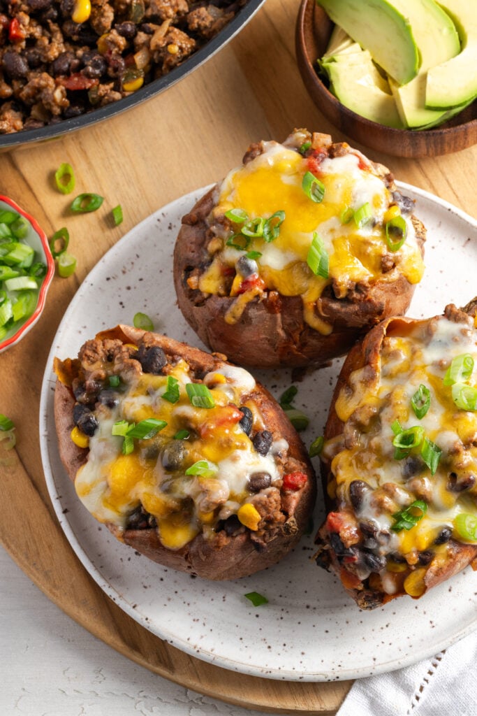 ground beef and black bean stuffed sweet potatoes topped with cheese and scallions on a white plate with a bowl of sliced avocado to the side.
