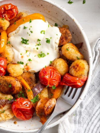 closeup shot of the right half of a bowl of baked gnocchi with a silver fork taking a bite with a roasted cherry tomato and golden brown gnocchi.