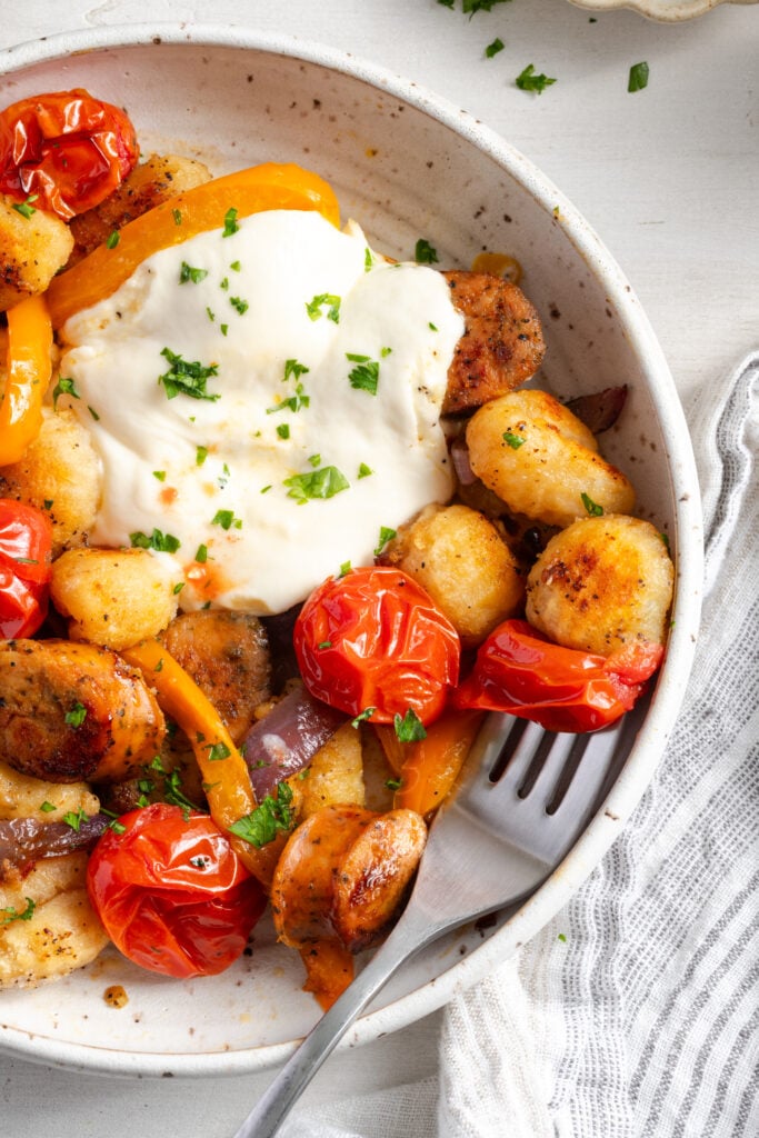 closeup shot of the right half of a bowl of baked gnocchi with a silver fork taking a bite with a roasted cherry tomato and golden brown gnocchi.