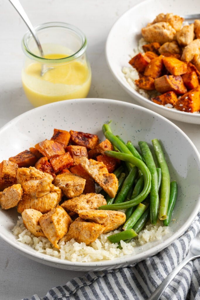 45 degree angle shot of a dinner table set with two chicken sweet potato bowls with green beans and cauliflower rice with a jar of garlicky citrus sauce to the side for drizzling.